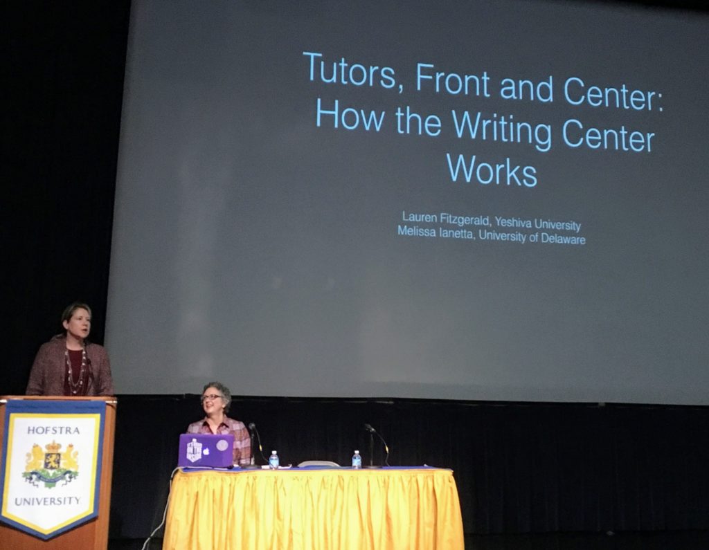 One woman standing at podium, the other seated at the dais.